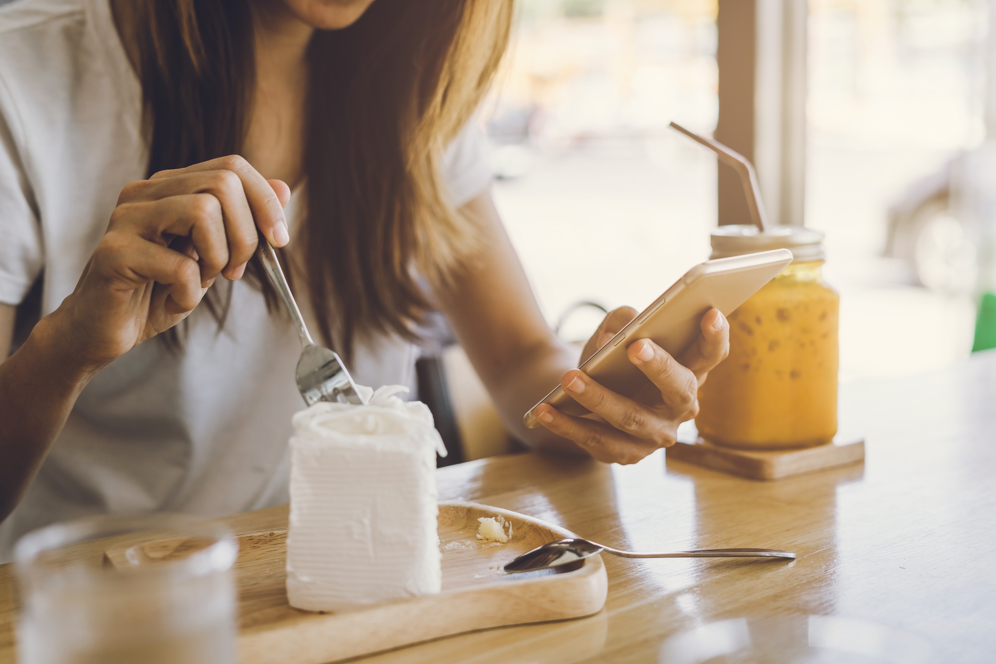 Young woman using smart phone and eating cake in cafe